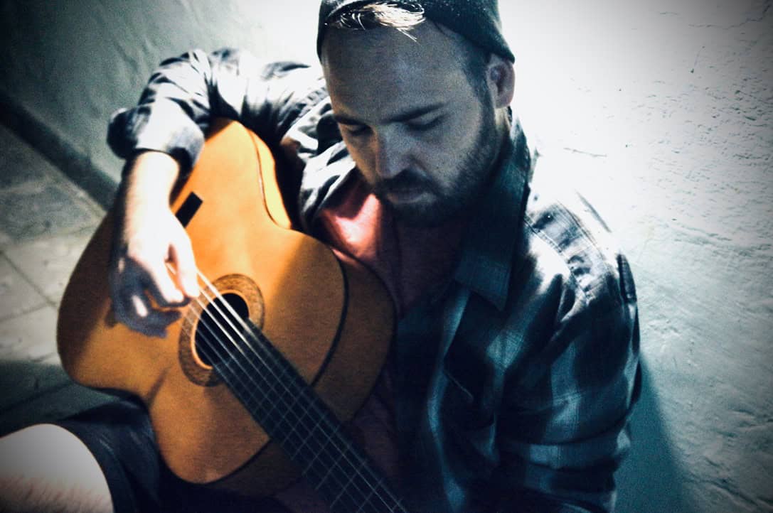 Chaz Chambers sits against a wall playing an acoustic guitar, wearing a plaid shirt and a backward cap.