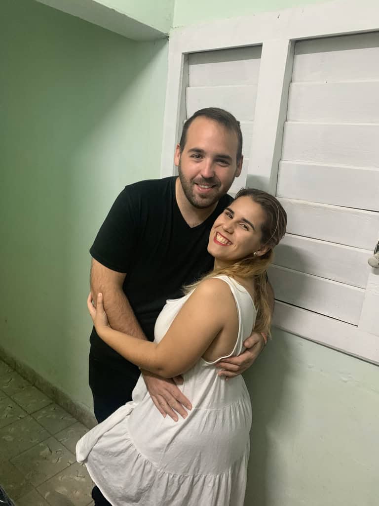 Chaz and Yami embrace and smile beside a white shuttered window and a pale green wall.