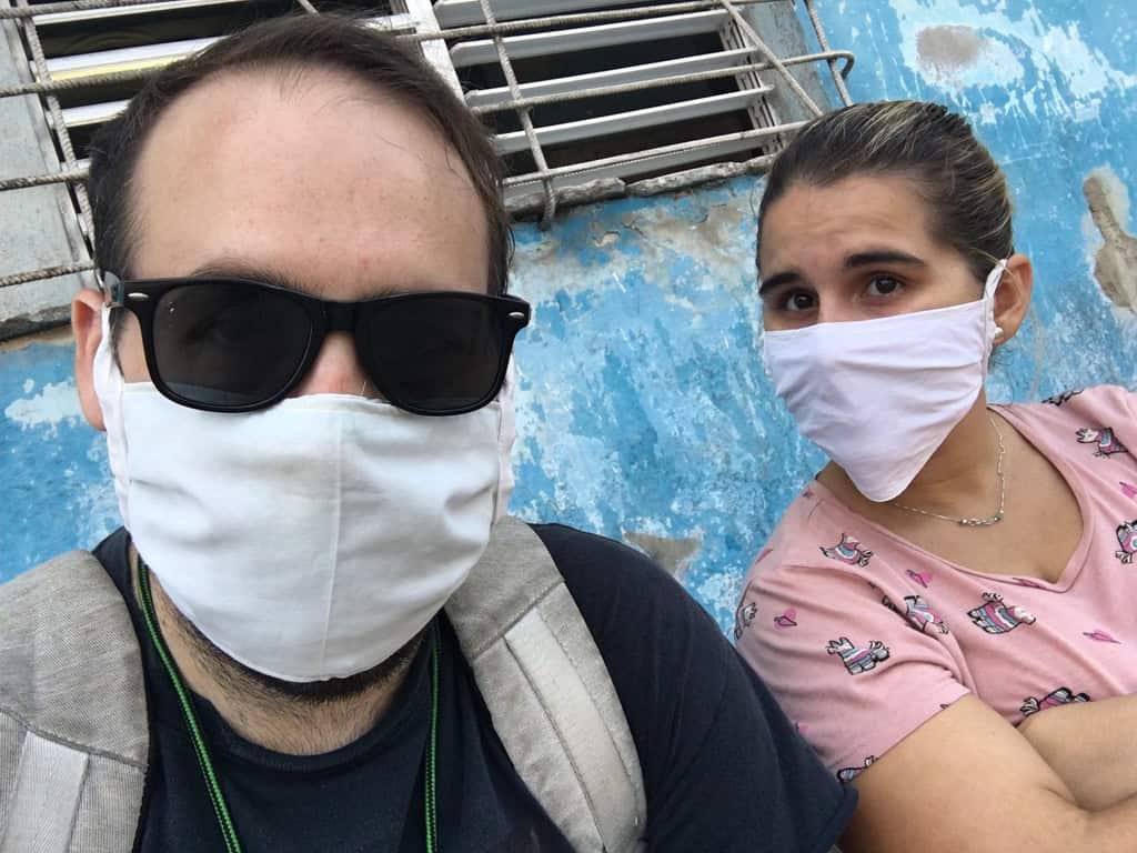 Chaz and Yami wear white cloth masks in front of a weathered blue wall and a barred, louvered window.