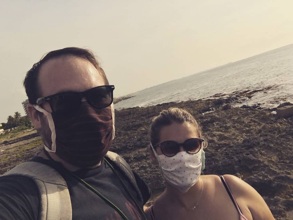 Chaz and Yami wear cloth face masks and sunglasses beside a rocky shoreline, with the sea behind them.