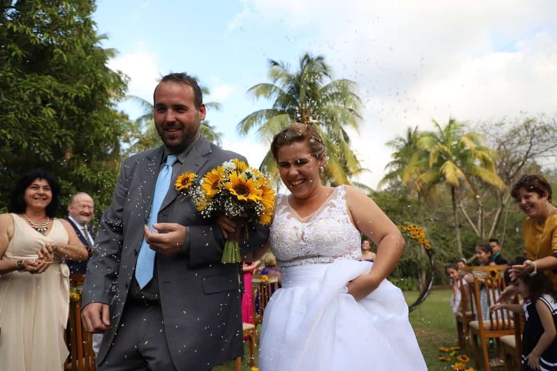 Chaz and Yami walk between wedding guests outdoors; he wears a gray suit and she carries a sunflower bouquet.