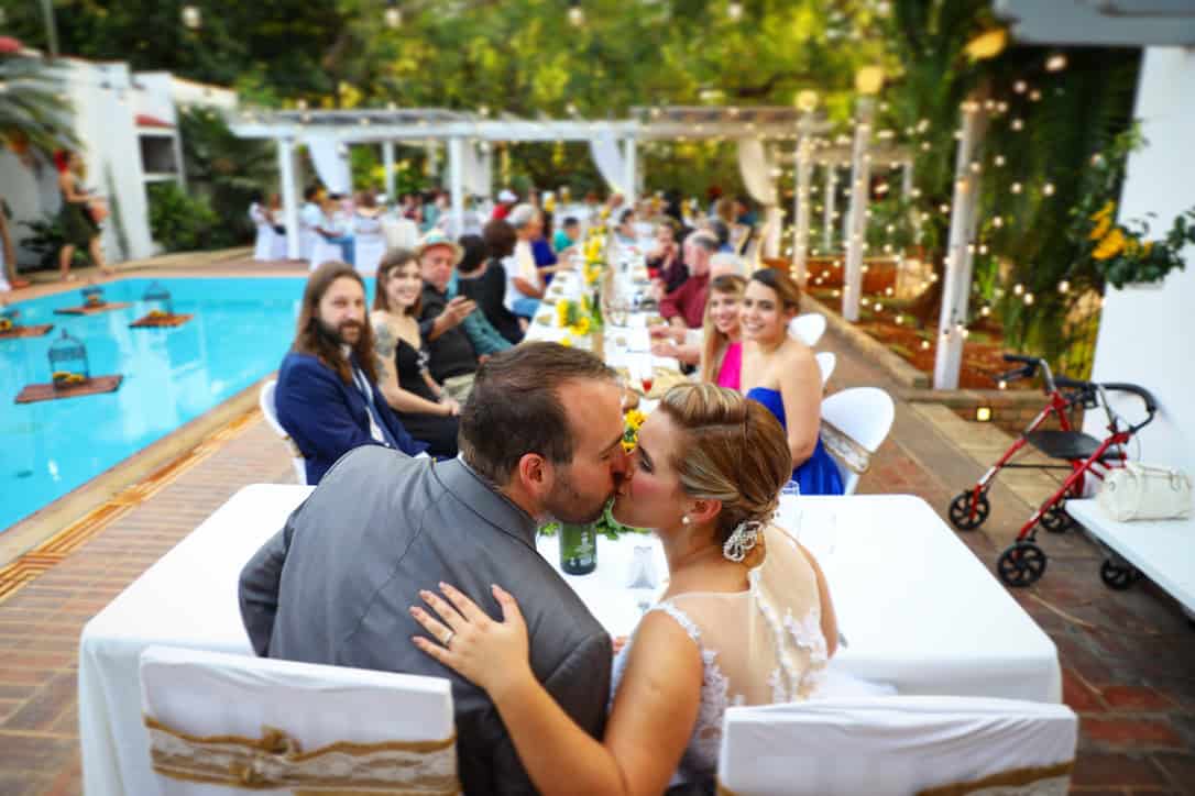 Chaz and Yami kiss at the end of a long wedding reception table beside a swimming pool, with guests behind them.