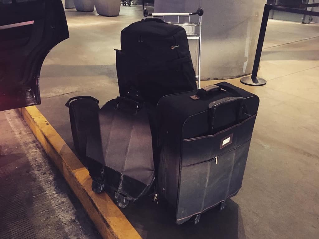 Black instrument cases, a rolling suitcase and a backpack beside a luggage cart at an airport curb.