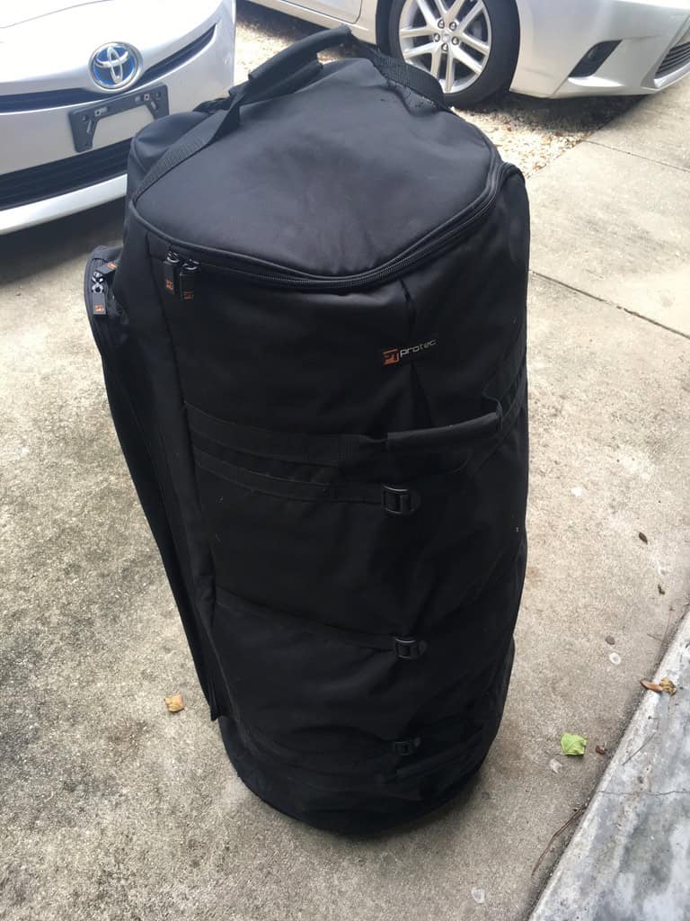 A tall black padded drum bag stands on a driveway beside two parked cars.